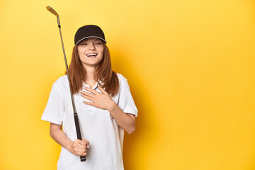 Redhead golfer with club and cap, studio shot laughs out loudly keeping hand on chest.