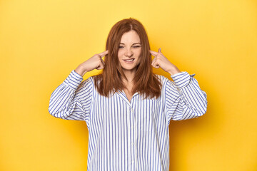 Business-dressed redhead, formal studio shot covering ears with fingers, stressed and desperate by...