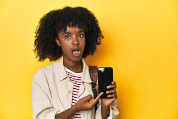 Teen girl showing smartphone, backpack on yellow background pointing to the side