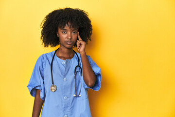 Young African-American nurse in studio with yellow background pointing temple with finger, thinking, focused on a task.
