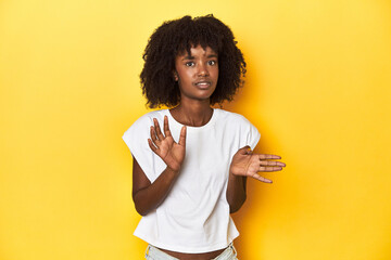 Teen girl in white tank top, yellow studio background rejecting someone showing a gesture of...