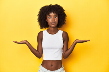 Teen girl in white tank top, yellow studio background confused and doubtful shrugging shoulders to hold a copy space. - obrazy, fototapety, plakaty