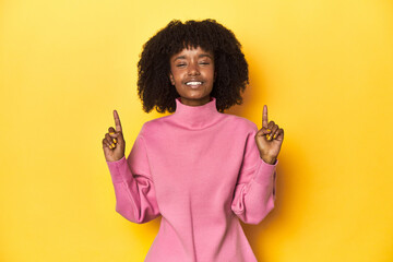 Teen girl in pink sweatshirt, yellow studio backdrop indicates with both fore fingers up showing a...