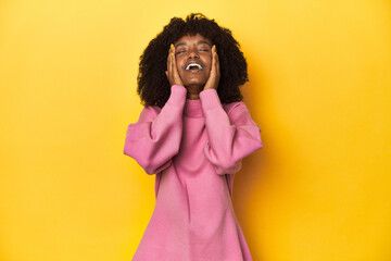 Teen girl in pink sweatshirt, yellow studio backdrop laughs joyfully keeping hands on head....