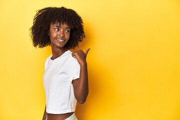 Teen girl in classic white T-shirt, yellow studio backdrop points with thumb finger away, laughing...
