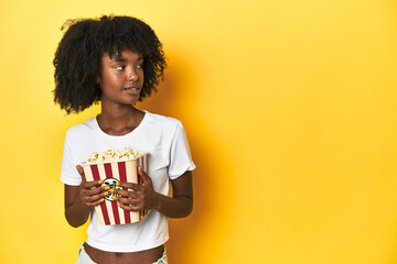 African-American teenage girl with popcorn, enjoying cinema concepts on yellow backdrop.