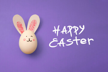 Chicken egg with cute rabbit face and bunny ears on lavender background. Food photo for Easter holiday. Greeting card. Happy Easter handwritten white text. Preparation for celebration. Family holiday