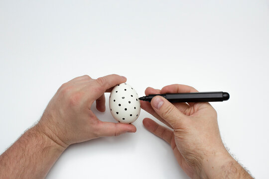 Male hairy hands with chicken egg on white background. Horizontal photo. Man draws polka dot pattern on eggshell with black marker. Concept of preparing for religious holiday, Happy Easter, creativity