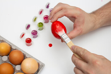Process of preparing for celebration of religious holiday. Happy Easter. Woman paints beige chicken egg with red paint on light white background. Brush in female hand. Finger with manicure. Egg box