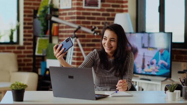 Laughing happy indian woman taking selfie with mobile phone while smiling at home office desk. Person in nice stylish modern house capturing photo using smartphone camera, camera B