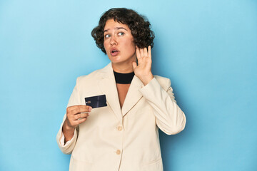 Young woman with credit card, elegant white blazer trying to listening a gossip.