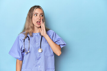 Young caucasian nurse in blue studio is saying a secret hot braking news and looking aside - obrazy, fototapety, plakaty