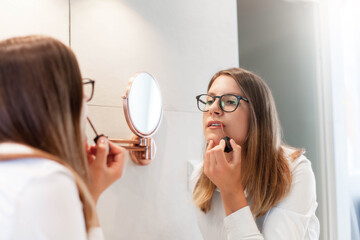 Young woman applying makeup in front of mirror in office restroom. Girl in eyeglasses using...