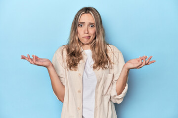 Blonde young caucasian woman in blue studio doubting and shrugging shoulders in questioning gesture.