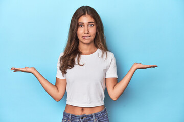 Teen girl in white T-shirt on a blue background confused and doubtful shrugging shoulders to hold a...