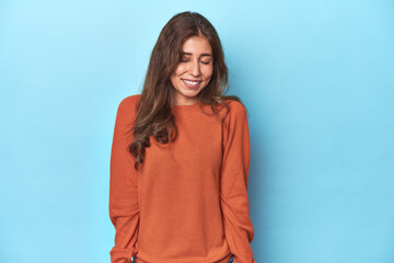 Teen girl in vibrant orange sweater on blue laughs and closes eyes, feels relaxed and happy.