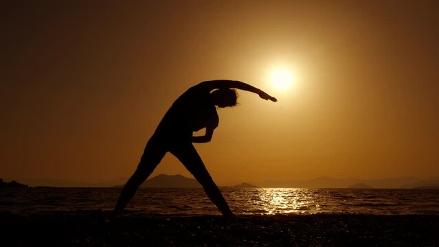 Female performing stretching yoga on shore. A view of woman silhouette stretching her body on the evening beach against sea horizon.