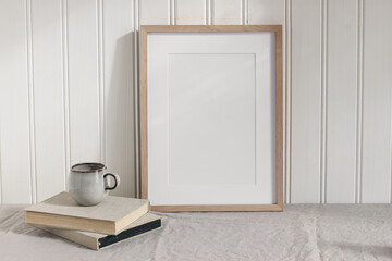 Blank vertical wooden picture frame mock up on linen table cloth. Modern interior. Cup of coffee on pile of vintage books. White wall background. Neutral Scandinavian home. Poster display template.