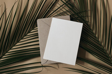 Summer tropical wedding stationery still life scene. Green date palm leaves. Blank greeting card, invitation mockup scene. Craft envelope. Beige table background. Flat lay, top view, no people.