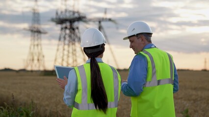 Electricians with tablet computer walk past power generation substation at countryside. Engineers study data on tablet controlling power plant against blurry transmission lines. Electricians in field