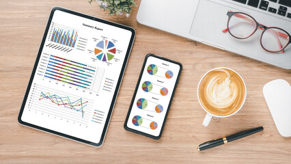 Business analytics concept, Tablet and smartphone displaying colorful charts and graphs on a wooden desk with a laptop, glasses, and a cup of coffee.