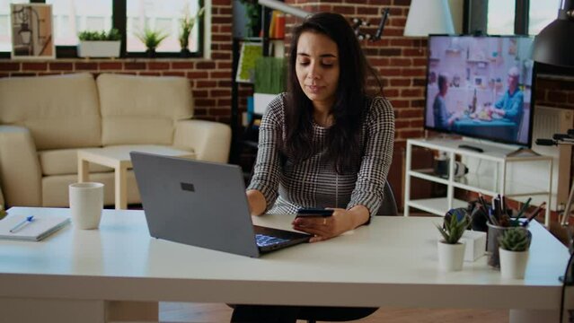 Happy woman stoked on doing online shopping, carefully typing credit card information on laptop. Indian person adding payment method on website while in cozy stylish warm home, camera A panning shot