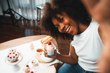Beautiful young African blogger presenting piece of cupcake in concept special cuisine with selfie on smartphone. Content creating of social media with favorite sweets bakery dish. Tastemaker. - obrazy, fototapety, plakaty