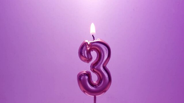 Full HD video. Birthday greeting card. Burning wax candle in form of the number 3. Purple paraffin candle melts from fire on lilac background. Drops of wax drip from heat. Concept of holiday, gift