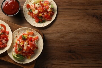 Delicious tacos with vegetables, lime and ketchup on wooden table, flat lay. Space for text