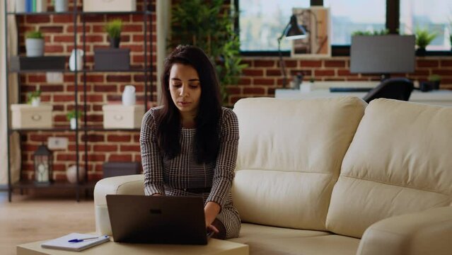 Indian teleworker sitting on couch, focused on finishing tasks in personal office. Remote worker concentrating on correctly inputting data on laptop, hurrying to complete project before deadline