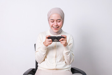 Excited young beautiful muslim woman with braces sitting on chair over white background playing games on mobile phone.