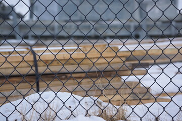 Fastened pile of timber planks on snowy ground, chainlink fence in foreground 