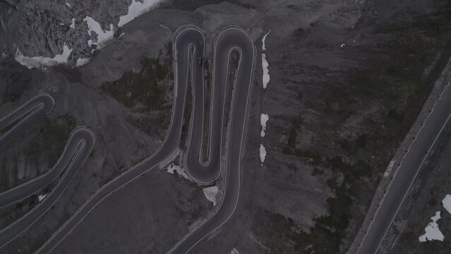 Top down drone shot of a car driving  down the hill on the Stelvio Pass Italy on a grey day with snow on the mountains on a twisty road and headlights on during sunset LOG