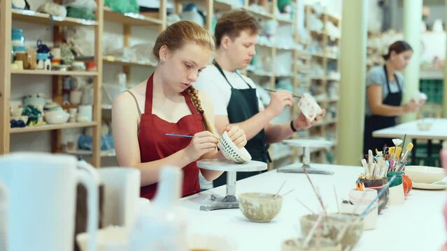 Young woman teacher showing teenage boy and girl how to paint ceramic cup