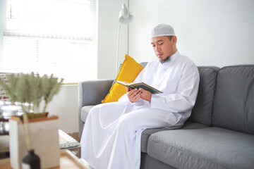 Asian muslim man sitting on sofa chair and reading the holy Quran