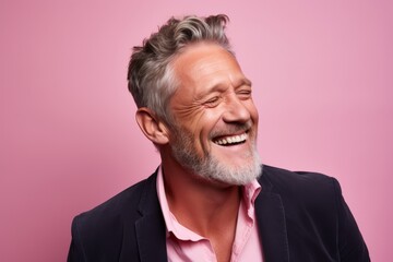 Handsome senior man with grey hair and beard laughing while standing against pink background