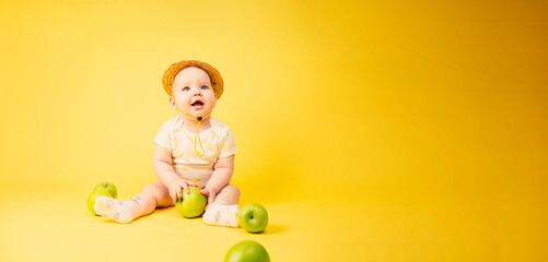 Beautiful baby girl in straw hat with green apples sitting in studio on yellow background.