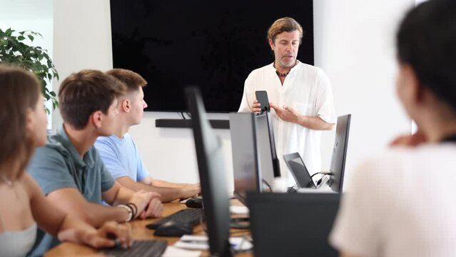 During lesson computer courses, male teacher holds mobile phone in hands and explains to listeners principle of operation.