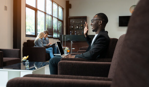 African businessman sitting in café or airport lounge using laptop and talking remote on video call. Young man sitting in armchair and waving hand in greeting gesture.