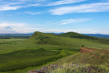 Natural background of hills with red rocks, green meadows and blue sky with clouds
