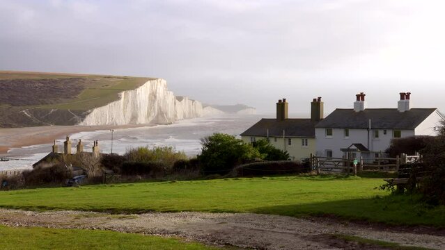 A row of buildings nestled on a land lot covered in lush green grass, with a Seven Sisters cliff in the background, under a sky dotted with fluffy clouds and windy ocean, England, Brighton, UK.