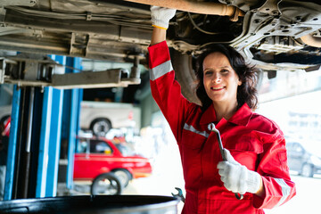 Portrait of a Caucasian female mechanic in a red uniform standing under the car bottom for inspecting in the garage. A woman smiling while holding a wrench. Car repair service. Vehicle maintenance