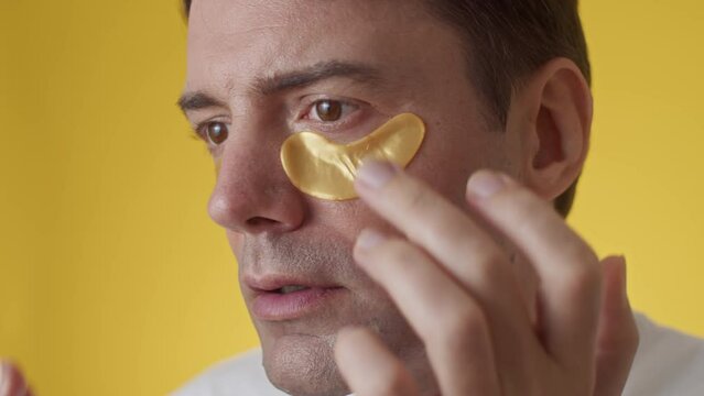 Big close up of middle-aged man wearing golden hydrogel under eye patches on yellow background