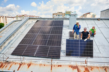 Workers building solar panel system on metal rooftop of house. Three men installers installing photovoltaic solar module outdoors. Alternative, green and renewable energy generation concept.
