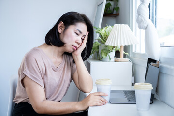 sleepless Asian woman feeling tired and fall asleep at home office with laptop and cup of coffee on desk