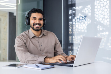 Carefree middle eastern guy in wireless headset typing on computer keyboard in modern office interior. Successful worker practising multitasking while listening to audiobook and answering emails.