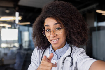 Diverse medic student in eyeglasses with bushy hairdo pointing at camera on blurred background....