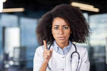 Serious african american doctor in white lab coat raising index finger with rigorous facial...