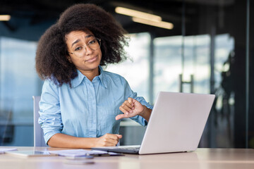 Upset black woman in blue shirt and eyewear making dislike gesture while working by desktop in office. Displeased female manager feeling bored during busy day and dreaming about changing position. - obrazy, fototapety, plakaty