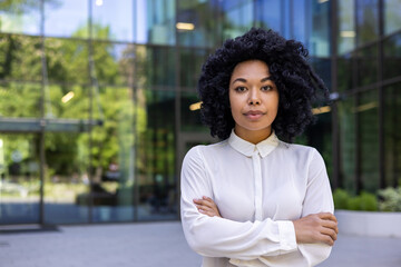 Serious black woman in white shirt standing with crossed arms on chest outside of glassy building...